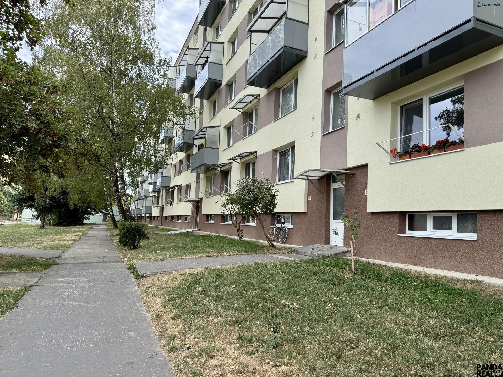 Bytový dům se zelení a chodníkem, nacházející se v Topoľčanech, obklopený stromy.