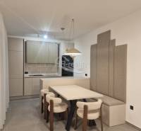 Jídelna v rekreačním apartmánu s kuchyní, světlými židlemi a šedými panely na stěně.