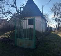 Malá chata obklopená stromy, oplocená zahrada, ideální pro rekreační pozemky.