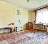 Pokoj s koberci, starším nábytkem a okny, praskliny na stěnách. Pozemky - bydlení.