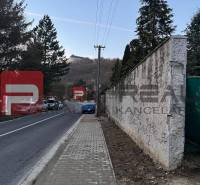Cesta s chodníkem vedoucím přes Pezinok u rodinného domu, v pozadí kopcovitá krajina.