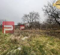 Pozemek s trávou a stromy na ulici Partizánska, v pozadí sklady a haly.