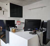 Kancelář se stolem, monitorem a poličkami, situovaná na ulici Partizánska u skladů.