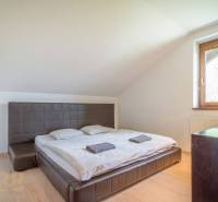 Ložnice v rekreačním apartmánu s manželskou postelí, oknem, dřevěným nábytkem a šikmými stropy.