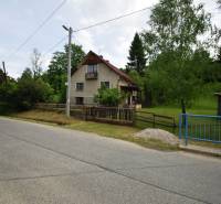 Rodinný dům ve Velké Lesné u cesty, obklopený zelení a plotem.