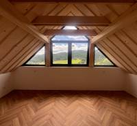 Podkrovní prostor v chatě s výhledem na hory a dřevěnými trámy.