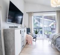 Světlá ložnice v rodinném domě s televizí, zrcadlem a balkonem.