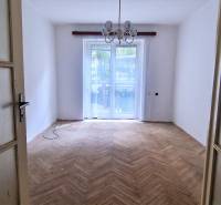 Prázdný pokoj s dřevěnou podlahou, velkým oknem a lustrem v 2-pokojovém bytě.