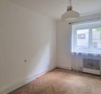 Pokoj s dřevěnou podlahou, oknem a bílými stěnami v 2-pokojovém bytě.