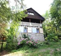 Chata v Holčíkovcích obklopená stromy a květinami, kamenné základy a bílý balkon.