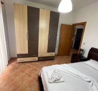 Světlá ložnice v rekreačním apartmánu s postelí, šatní skříní a dlaždicovou podlahou.