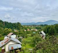 Venkovská chalupa v Mojši obklopená zelení a kopci pod modrou oblohou.