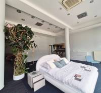 Postel s polštáři a rostlinami v obchodních prostorách, vedle noční stolek a křeslo.