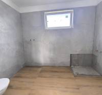 Koupelna v rodinném domě s dlažbou, připravená k instalaci, světlo přes malé okno.