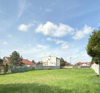 Pozemek k bydlení ve Sládkovičově, tráva, ploty, stromy, budovy v pozadí, oblačná obloha.
