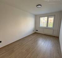 Prázdný pokoj s dřevěnou podlahou v 3-pokojovém bytě, velké okno a radiátor pod ním.