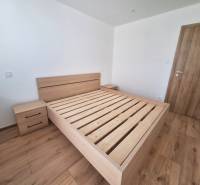 Ložnice s dřevěnou postelí v dvoupokojovém bytě, bez matrace a s nočním stolkem.