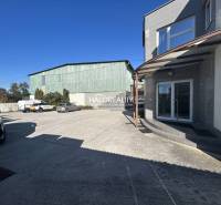 Exteriér výrobního prostoru s parkovištěm a několika zaparkovanými auty pod modrou oblohou.