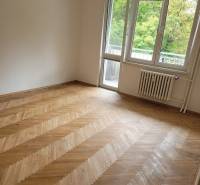Pokoj ve 3-pokojovém bytě s dřevěnou podlahou a balkonovými dveřmi.