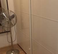Šatník v 2-pokojovém bytě s bílými posuvnými skříněmi a stojanovým ventilátorem.