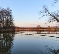 Jezero s můstkem, obklopené holými stromy a v pozadí teplé barvy západu slunce.