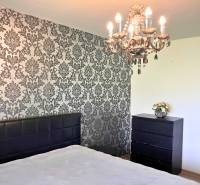 Ložnice s lustrem a tapetovou stěnou v rodinném domě. Podlaha s dřevěným dekorem.