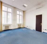 Interiér kanceláře s modrým kobercem, bílými stěnami a velkými okny.