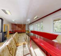 Pokoj v chatě s kachlovými kamny, pohovkami, dřevěnými stoly a výhledem na les.