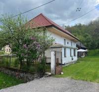 Venkovská chata obklopená zelení, s rustikální střechou, kvetoucí zahradou a dřevěným altánkem.