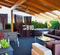 Terasa rodinného domu s posezením a grilem, částečně krytá s dřevěnou střechou.