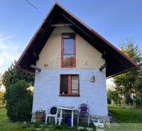 Chata v Sološnici se strmou střechou, okna, příroda a plastové židle.