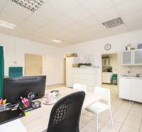 Kancelář s nábytkem, počítačem a kuchyňkou, na podlaze jsou dlaždice.