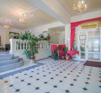 Hotelová recepce s rostlinami a lustry, dlažba s geometrickým vzorem.