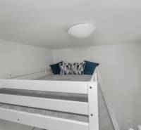 Postel na vyvýšeném místě v garsonce s polštáři a modrým závěsem.