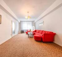 Obývací pokoj se světlou stěnou, červenou sedačkou a kobercem ve 4pokojovém bytě.