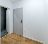 Chodba se šedými dveřmi, koupelna s tmavými obklady a podlahou s dřevěným dekorem.