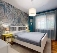 Ložnice v rodinném domě s palmami na tapetě, podlahou s dřevěným dekorem a bílým nábytkem.