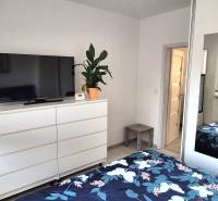 Ložnice v dvoupokojovém bytě s bílým nábytkem, zrcadlem a televizí na komodě.