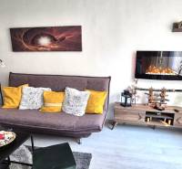 Obývací pokoj v 2-pokojovém bytě s pohovkou, policí a elektrickým krbem.