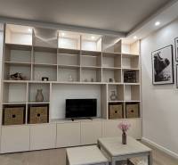 Úložná stěna s policemi a dekoracemi v 2-pokojovém bytě, vedle stůl s květinami.