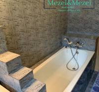 Koupelna se schůdky a vanou obklopená šedými obklady v 2pokojovém bytě.