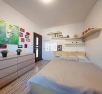 Ložnice v rodinném domě s postelí, poličkami, obrazy a podlahou s dřevěným dekorem.