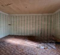 Pokoj v rodinném domě s poškozeným stropem, tapetami a podlahou s dřevěným dekorem.