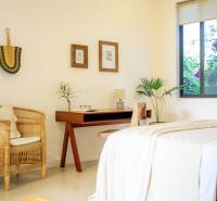 Světlá ložnice v rodinném domě s ratanovým křeslem a dřevěným stolem.