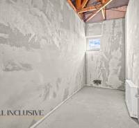 Nedokončená místnost, na stěnách šedá omítka, s malým oknem a viditelnou střešní konstrukcí.