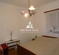 Ložnice v rodinném domě s postelí, stolkem a lustrem, na stěně je okno se záclonou.