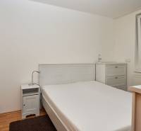 Ložnice v dvoupokojovém bytě s manželskou postelí, nočním stolkem a komodou.