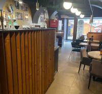 Interiér gastro prostorů s dřevěným barem a stoly. Vidět je i dekorativní osvětlení.