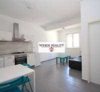 Obývací pokoj v dvoupokojovém bytě s kuchyňským koutem, jednoduchým zařízením a oknem.