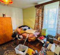 Pokoj v rodinném domě s nepořádkem, starým nábytkem a výhledem z okna.